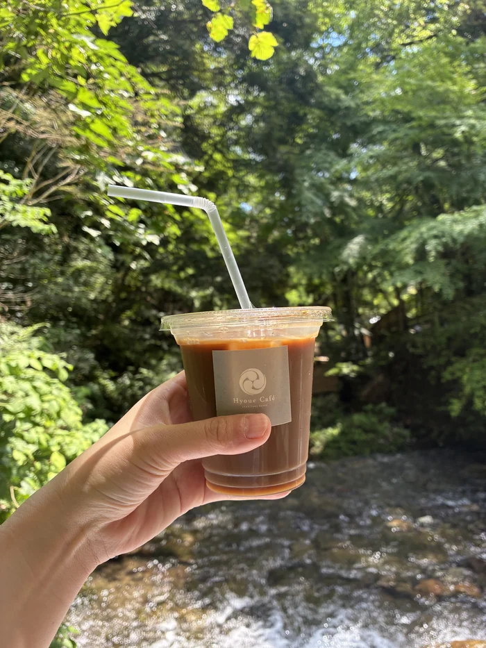 Hyoue Cafeのロゴ入りカップのアイスコーヒーを、川を背に手に持つ様子