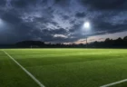 ナイター照明に照らされた無人のサッカーピッチ、夕暮れの曇天を広角でとらえた情景