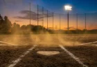 夕暮れのナイター照明と砂ぼこりが舞う本塁付近の写実的な情景。人物・文字なし