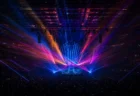 大型アリーナのコンサート会場、暗いステージを彩る色とりどりのスポットライトとレーザー光線、遠景の客席シルエット