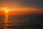 夕暮れの穏やかな海と水平線。遠景に古代遺跡のシルエットが浮かび、金色の光が水面を照らす幻想的な情景