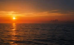 夕暮れの穏やかな海と水平線。遠景に古代遺跡のシルエットが浮かび、金色の光が水面を照らす幻想的な情景