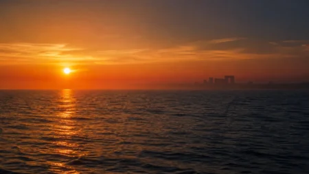 夕暮れの穏やかな海と水平線。遠景に古代遺跡のシルエットが浮かび、金色の光が水面を照らす幻想的な情景