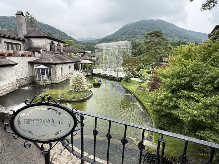 箱根ガラスの森美術館の看板越しに池とヴェネチア風の建物と光の回廊と山並みを見渡す景色