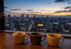 東京の高層階から街の夜景を望むカフェの窓辺に3つのコーヒーカップが並ぶ写実的な情景。人物・文字なし