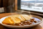 木製テーブルに置かれた、とろけるチーズソースがかかったオムレツとカレーの一皿。湯気が立ち、窓の外に雪景色が見える温かみのある情景