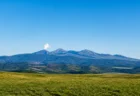 青空の下、緑の草原の向こうに控えめな噴煙を上げる火山の山並み