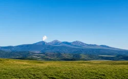 青空の下、緑の草原の向こうに控えめな噴煙を上げる火山の山並み