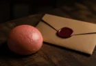 木製テーブルに置かれた赤みがかったピンク色の饅頭と、封蝋付きの茶封筒が並ぶスパイ映画風の写実的な情景