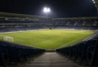 夜のサッカースタジアム、無人のスタンド席の通路から見下ろす芝生のピッチ、ナイター照明が柔らかく灯る情景