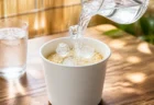 湯気の出ていない無地の白いカップ麺に氷と冷たい水を注ぐ夏の情景、隣に結露したグラス
