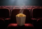 暗い映画館の赤いベルベット座席に置かれたポップコーン容器から立ち上る蒸気、スクリーンからの淡い光