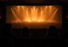 暗い映画館の客席から見た大スクリーンに、暖色のスポットライトが照らすコンサート会場の遠景が映し出されている情景、観客の後ろ姿シルエット
