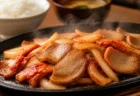 鉄板の上で湯気を立てながら豚肉とキムチを炒め焼きにしている情景、奥にご飯とみそ汁がぼんやり写る定食屋の食卓