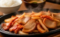 鉄板の上で湯気を立てながら豚肉とキムチを炒め焼きにしている情景、奥にご飯とみそ汁がぼんやり写る定食屋の食卓