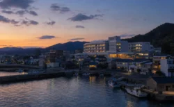 夕暮れ時、海沿いの港町に建つ病院と街並みを望む写実的情景
