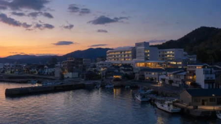 夕暮れ時、海沿いの港町に建つ病院と街並みを望む写実的情景