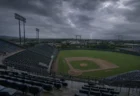 夕暮れの雨雲と遠方の稲光の下、無人の野球場スタンド席とグラウンドを見下ろす情景