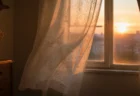 夕暮れの柔らかなオレンジ光が差すレースカーテンの揺れる家庭的な窓辺のイメージ