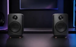 暗いゲーミングデスク上に置かれた左右一対のモダンなワイヤレススピーカーを、淡い間接照明の中で写した写実的な情景