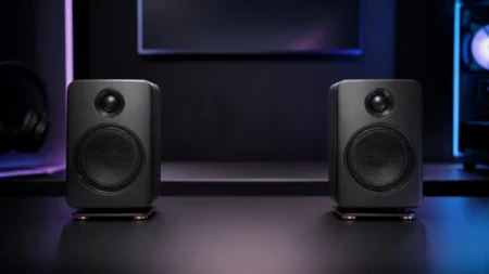 暗いゲーミングデスク上に置かれた左右一対のモダンなワイヤレススピーカーを、淡い間接照明の中で写した写実的な情景