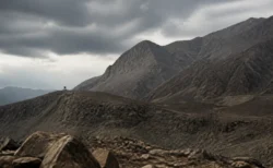 険しい岩山と曇り空、遠くに小さな監視塔のシルエットが見える荒涼とした山岳地帯の情景