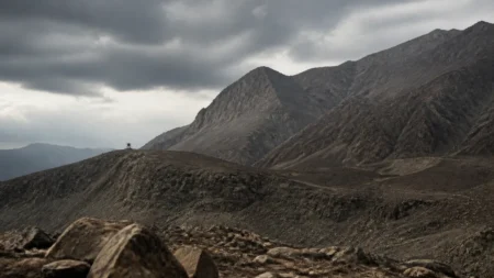 険しい岩山と曇り空、遠くに小さな監視塔のシルエットが見える荒涼とした山岳地帯の情景