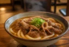 湯気の立つ牛肉うどんが木製テーブルの丼に盛られたうどん専門店店内のイメージ