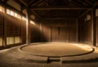 早朝の相撲部屋の稽古場、砂がならされた無人の土俵に朝の光が差し込む写実的情景