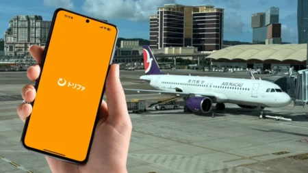 マカオ国際空港の駐機場を背景に、トリファのアプリを起動したスマートフォンを手に持っている様子