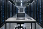 データセンターのサーバーラックが並ぶ通路に、閉じたノートパソコンと無地の入館証が置かれた事務机が1台だけある情景
