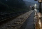 夜明け前の無人のローカル線ホームに激しい雨が降り注ぎ、線路脇が増水している様子
