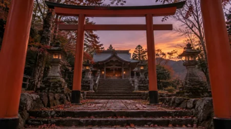 夕暮れの神社の境内。朱色の鳥居越しに見える木造の社殿と石灯籠、落ち葉の積もった石段