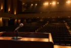 演説会場の演台に置かれたマイクと、客席に向けて照らされたライトの写実的な風景