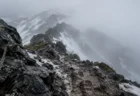 濃い霧が垂れこめる北海道の山の稜線。岩場の登山用ロープとぬかるんだ登山道の足跡