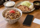 木のカウンターに置かれた牛丼と定食のプレート、その手前のスマートフォンと無地の白いカード