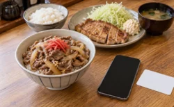 木のカウンターに置かれた牛丼と定食のプレート、その手前のスマートフォンと無地の白いカード