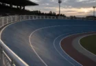 夕暮れの無人の競輪場のバンク。青い走路と白いライン、外柵のフェンスが写る
