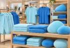 明るい衣料品店の売り場。青や水色のパーカー、Tシャツ、ブランケット、丸いクッションが並んでいる
