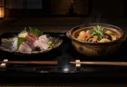 黒い漆の膳に向かい合わせに置かれた刺身の盛り合わせと土鍋の煮物。暗い和室の柔らかな光