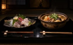 黒い漆の膳に向かい合わせに置かれた刺身の盛り合わせと土鍋の煮物。暗い和室の柔らかな光