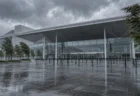 雨に濡れた大型展示会場の屋外エントランス広場。閉じたガラス扉と無人のベルトパーティション、低い雨雲