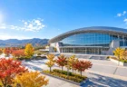 秋晴れの空の下に建つ屋内アリーナと紅葉した木々（イメージ）