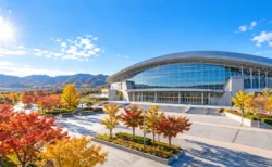 秋晴れの空の下に建つ屋内アリーナと紅葉した木々（イメージ）