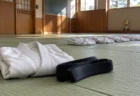 午後の光が差し込む柔道場の畳に置かれた白い柔道着と黒帯、奥に並ぶ子ども用の柔道着と優勝カップ