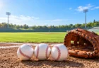 晴れた野球場の内野の土に並んだ3つの硬式野球ボールと茶色のグラブ
