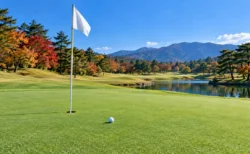 秋の紅葉に囲まれたゴルフ場のグリーンとピンフラッグ、白いボール