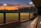 夕暮れのナイター照明がともる野球場を、誰もいないダッグアウトのベンチ越しに望む風景