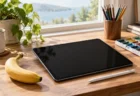 明るい窓辺の木のデスクに置かれた画面消灯のタブレットとスタイラスペン、色鉛筆、絵の具のパレットとバナナ