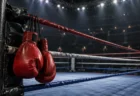 照明に照らされた無人のボクシングリングと、コーナーポストに掛けられた赤いボクシンググローブ